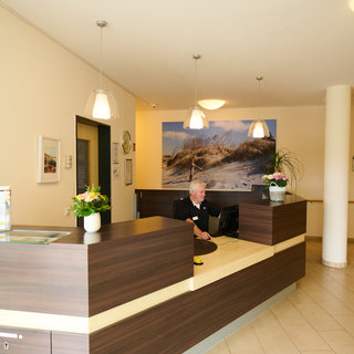 Ein Empfangsbereich mit einer Rezeption aus Holz. Eine Person sitzt am Computer und begrüßt Gäste. An der Wand hängt ein Bild von Dünen. Die Beleuchtung ist hell, und es gibt einige Pflanzen auf dem Empfangstresen. Der Raum wirkt freundlich und einladend.
