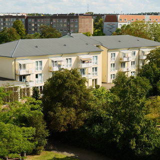Ein mehrstöckiges Wohngebäude mit Balkonen ist von Bäumen und Grünanlagen umgeben. Im Hintergrund sind weitere Wohnblocks sichtbar. Der Himmel ist klar und es ist Tag.