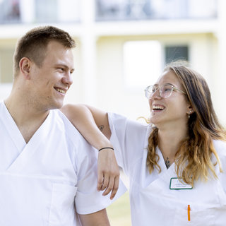 Zwei junge Menschen in weißen Arbeitskleidung lächeln sich an, stehend auf einer grünen Wiese. Der Mann hat kurze Haare und der Frau trägt eine Brille. Sie wirken freundlich und entspannt, während sie in einer outdoor-Umgebung miteinander interagieren.