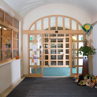 Das Bild zeigt einen einladenden Eingangsbereich mit einer großen Glastür und Holzelementen. Links befindet sich ein Empfangstresen, während dekorative Figuren und Pflanzen den Boden schmücken. Der Raum wirkt hell und freundlich, mit einem Teppich und grünen Akzenten.