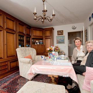 Zwei Frauen sitzen an einem Tisch in einem gemütlichen Wohnzimmer. Der Raum ist mit holzfarbenen Möbeln eingerichtet, und auf dem Tisch stehen Blumen und Dokumente. Die Stimmung ist freundlich und einladend.