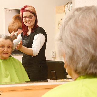 Eine ältere Frau mit grauen Haaren sitzt in einem Friseurstuhl und trägt einen grünen Umhang. Eine Friseurin mit rotem Haar steht hinter ihr und schneidet ihr Haar. Die Frau lächelt und schaut in den Spiegel.