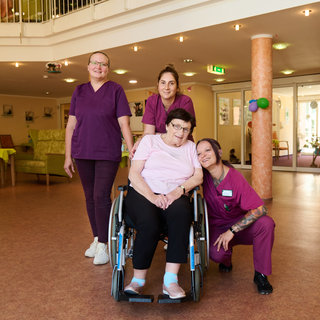 Vier Personen stehen in einem Gemeinschaftsraum. Eine Frau sitzt im Rollstuhl, umgeben von drei Pflegekräften, die freundliche Ausdrücke zeigen. Der Raum wirkt hell und einladend, mit Sitzgelegenheiten und dekorativen Elementen im Hintergrund.