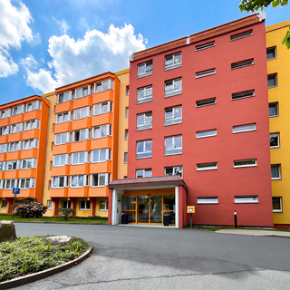 Bunt gestrichene Wohngebäude mit orangefarbenen und roten Fassaden unter einem blauen Himmel mit Wolken. Im Vordergrund eine Zufahrt mit einem kleinen Rasen und einigen Bäumen.