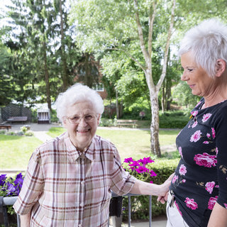 Eine ältere Frau mit Brille und kariertem Hemd steht lächelnd auf einem Balkon. Neben ihr steht eine jüngere Frau mit kurzem, blondem Haar und einem blumigen Oberteil. Im Hintergrund sind grüne Bäume und Blumen sichtbar, was eine freundliche und einladende Atmosphäre schafft.