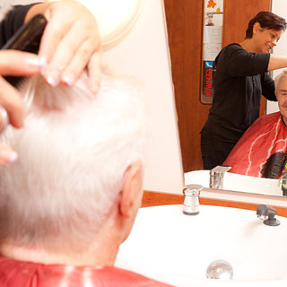Ein Friseur schneidet die Haare eines älteren Mannes, der in einem Friseurstuhl sitzt und ein rotes Umhang trägt. Im Hintergrund ist eine Frau zu sehen, die lächelt und das Haar schneidet. Im Spiegel sind die beiden Personen sichtbar.