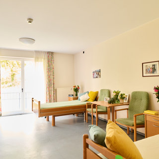 Ein heller, freundlicher Raum mit zwei Betten, einem Tisch, Stühlen und Blumenarrangements. Große Fenster mit Vorhängen lassen Tageslicht herein. An der Wand hängen Bilder. Der Raum wirkt einladend und gemütlich.