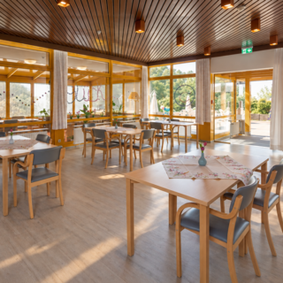 Ein helles, einladendes Restaurant mit mehreren Holztafeln und Stühlen. Große Fenster bieten Blick auf den Außenbereich. Die Räume wirken freundlich und gut beleuchtet, mit Pflanzen und dekorativen Elementen.