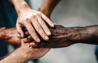 Mehrere Hände verschiedener Hauttöne liegen übereinander in einer verbandelten Formation. Die Bildkomposition symbolisiert Zusammenarbeit, Einheit und Solidarität zwischen unterschiedlichen Menschen.