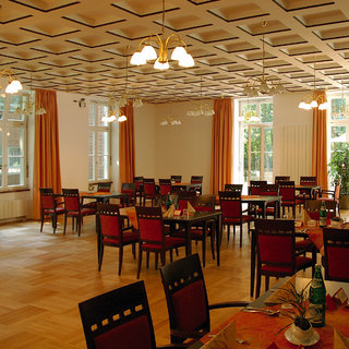 Ein heller Speisesaal mit Holzfußboden und großen Fenstern. Tische sind mit roten Stühlen umgeben, und einige Tische sind gedeckt. Im Hintergrund ist eine Person zu sehen. Der Raum hat eine gemütliche Atmosphäre mit bunten Vorhängen und warmem Licht.