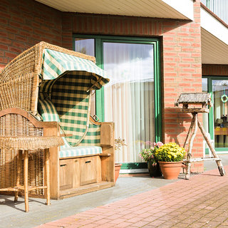 Ein Strandkorb aus Korbgeflecht mit grünen und weißen Streifen steht auf einem gepflasterten Gehweg vor einem Gebäude. Neben dem Korb befindet sich ein kleiner Tisch, und im Hintergrund ist ein farbenfroher Blumentopf zu sehen.