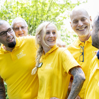 Eine Gruppe von sechs Personen in gelben Polo-Shirts steht zusammen im Freien. Sie lächeln und unterhalten sich freundlich. Die Atmosphäre ist fröhlich und einladend, was auf Teamgeist und Zusammenarbeit hinweist. Im Hintergrund sind Bäume und grünes Laub sichtbar.