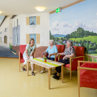 Drei Personen, zwei ältere Frauen und eine jüngere Frau, sitzen in einem gemütlichen Raum mit roten Sesseln und einem Tisch. Im Hintergrund sind Wandbilder von einer Landschaft und einer Straße zu sehen, die eine einladende Atmosphäre schaffen.