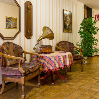 Ein Flur mit zwei gepolsterten Stühlen und einem kleinen Tisch. Auf dem Tisch steht ein Grammophon, umgeben von einer karierten Tischdecke. An den Wänden hängen Spiegel und Bilder. Eine Pflanze steht in der Ecke, und der Boden ist mit Fliesen ausgelegt.