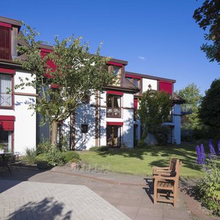 Ein modern gestaltetes Gebäude mit rotem Fensterladen und mehreren Fenstern umgeben von einem grünen Garten. Im Vordergrund befinden sich Pflanzen und ein Holzstuhl. Der Himmel ist blau und es gibt einige Bäume und Sträucher im Garten.