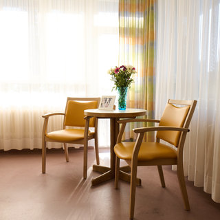 Ein heller Raum mit großen Fenstern und sanften Vorhängen. In der Mitte stehen ein runder Tisch und zwei gelbe Stühle. Auf dem Tisch befindet sich eine Vase mit Blumen und ein Bilderrahmen. Der Raum hat einen ruhigen und einladenden Charakter.