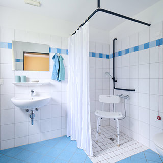 Das Bild zeigt ein Badezimmer mit weißen Fliesen und blauen Akzenten. Auf der linken Seite befindet sich ein Waschbecken mit Spiegel, in der Mitte eine Duschkabine mit Vorhang und rechts eine Toilette. Es gibt auch einen Duschstuhl und Haltegriffe für mehr Sicherheit.