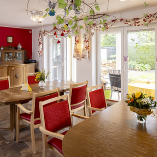 Ein heller Essbereich mit einem großen Holztisch und Stühlen in Rot. Auf dem Tisch steht eine Blumenvase mit bunten Blumen. Die Wände sind rot und weiß, und große Fenster bieten einen Blick nach draußen in den Garten.