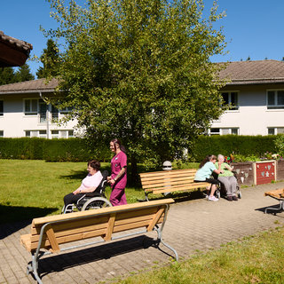 Eine Pflegerin schiebt eine Rollstuhlfahrerin durch einen Aufenthaltbereich im Freien, umgeben von Bänken und einem großen Baum. Im Hintergrund sitzen zwei weitere Personen auf einer Bank. Die Szene spielt in einer hellen, einladenden Umgebung mit einem klaren blauen Himmel.