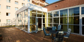 Ein helles, glasüberdachtes Terrassenzimmer mit Blick auf einen Außenbereich. Vier gepolsterte Stühle und ein Tisch stehen auf dem gepflasterten Boden. Im Hintergrund ist ein mehrstöckiges Wohngebäude zu sehen, das von Sonnenlicht beleuchtet wird.