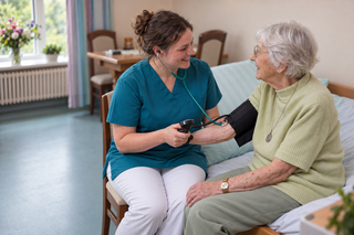 Eine Krankenschwester misst der älteren Patientin den Blutdruck. Beide lächeln und sitzen auf einem Bett in einem hellen Raum. Im Hintergrund sind ein Tisch und Blumen zu sehen.