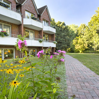 Eine Wohnanlage mit zwei Etagen, die Balkone mit Blumenarrangements haben. Im Vordergrund blühen gelbe und rosa Blumen. Ein gepflasterter Weg führt durch eine grüne Wiese, die von Bäumen umgeben ist, im Hintergrund ist ein Pavillon sichtbar.