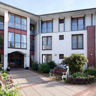 Das Bild zeigt das Eingangsgebäude einer Einrichtung mit mehreren Fenstern und einem einladenden Eingangsbereich. Vor dem Gebäude befinden sich bepflanzte Beete und Sitzbänke, die den Außenbereich gestalten. Die Architektur ist modern und freundlich.