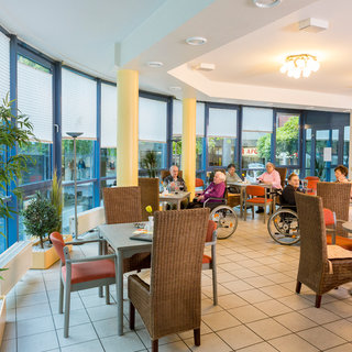 Das Bild zeigt einen hellen Speisesaal in einem Seniorenheim. In der Mitte sitzen mehrere ältere Menschen an Tischen, einige im Rollstuhl. Große Fenster mit natürlichem Licht schaffen eine freundliche Atmosphäre, während Pflanzen im Raum das Ambiente auflockern.