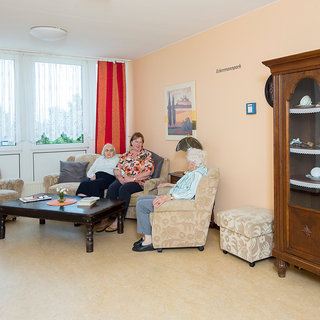 Drei Frauen sitzen in einem gemütlichen Aufenthaltsraum. Sie unterhalten sich, während sie auf einem beigen Sofa und einem Sessel platzgenommen haben. Neben ihnen steht ein Tisch mit einigen Büchern. Im Hintergrund sind große Fenster, die viel Tageslicht hereinlassen.