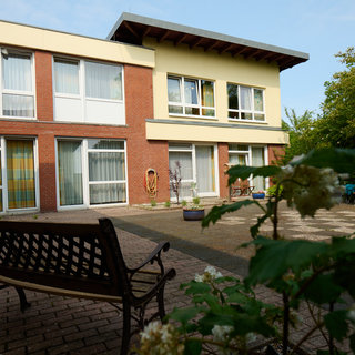 Eine Ansicht eines Wohngebäudes mit mehreren Fenstern, umgeben von einem gepflegten Garten. Zu sehen sind zwei Personen, die an einem Tisch sitzen, sowie eine Person im Rollstuhl. Die Atmosphäre wirkt freundlich und einladend.