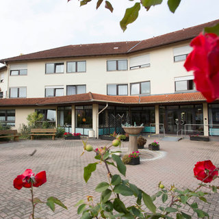 Ein seniorengerechtes Wohnheim mit mehreren Geschossen und großen Fenstern. Der Eingangsbereich ist mit einem Brunnen und Blumenrabatten gestaltet. Im Vordergrund sind rote Rosen zu sehen, die den einladenden Außenbereich schmücken.