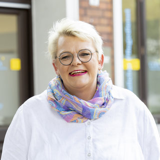 Eine fröhliche Frau mit blonden Haaren und einer bunten, knotenförmig gebundenen Schal lächelt in die Kamera. Sie trägt ein weißes Oberteil und steht vor einem Gebäude mit großen Fenstern. Die Atmosphäre wirkt freundlich und einladend.