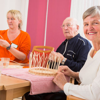 Drei Personen sitzen an einem Tisch und arbeiten an einem Bastelprojekt. Eine ältere Frau lächelt, während sie einen Korb aus Holzstreifen zusammenstellt. Neben ihr sitzt ein älterer Mann, der ebenfalls bastelt, und eine Frau im orangen Shirt erklärt das Vorgehen.