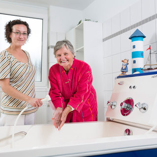 Eine Pflegerin steht neben einer älteren Frau, die an einem Waschbecken sitzt. Die Pflegerin hilft der Frau beim Händewaschen. Beide lächeln. Im Hintergrund ist eine Badewanne und ein dekorativer Leuchtturm zu sehen.