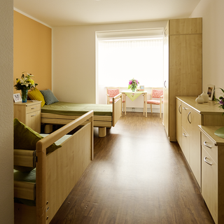 Ein heller Raum mit zwei Betten, einem Fenster und einer kleinen Sitzecke. An einer Wand stehen zwei Stühle und ein Tisch mit Blumen. Holzmöbel und ein sanfter Farbton schaffen eine einladende Atmosphäre. Der Boden ist aus Holz.