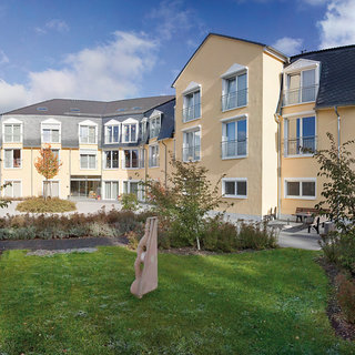 Das Bild zeigt eine moderne Wohnanlage mit hellen, gelben Gebäuden und großen Fenstern. Im Vordergrund befindet sich ein gepflegter Garten mit grünen Rasenflächen und kleinen Bäumen. Ein Holzskulptur steht im Garten. Die Atmosphäre wirkt einladend und freundlich.