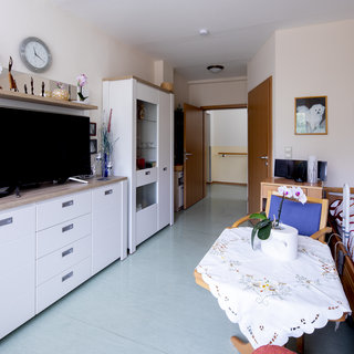 Ein modernes Zimmer mit einem Fernseher auf einer weißen Kommode, einem gepolsterten Bett mit rotem Muster und einem Tisch, der mit einer Tischdecke und einer Blumenvase dekoriert ist. Der Raum hat helle Wände und einen warmen, einladenden Einfluss.