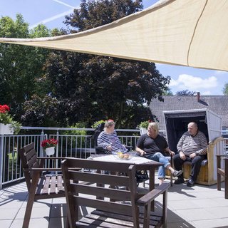 Drei Personen sitzen entspannt auf einer Terrasse, umgeben von Pflanzen. Ein Sonnensegel bietet Schatten. Die Szene vermittelt eine gemütliche Atmosphäre an einem sonnigen Tag.