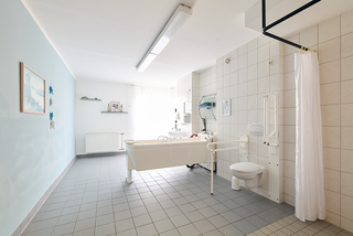 Ein barrierefreies Badezimmer mit einem großen, weißen Badewanne, einem Toilettenbereich und einem Duschvorhang. Helle Fliesen und eine blaue Wand schaffen eine freundliche Atmosphäre. Es gibt Fenster für natürliches Licht und einige Dekorationsobjekte an den Wänden.