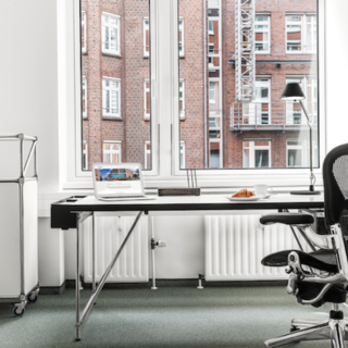 Ein moderner Arbeitsplatz mit einem Schreibtisch, einem ergonomischen Stuhl und einem Laptop. Neben dem Schreibtisch steht ein Rollwagen mit einem Getränk. Große Fenster bieten Blick auf ein Backsteingebäude. Der Boden ist mit grünem Teppich ausgelegt.