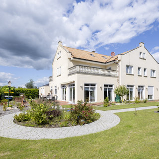 Ein helles zweigeschossiges Haus mit einem Balkon und mehreren Fenstern steht in einem gepflegten Garten. Der Garten ist mit bunten Pflanzen und einem geschwungenen Weg aus Pflastersteinen gestaltet. Im Hintergrund sind große, grüne Hecken und einige weitere Häuser zu sehen.