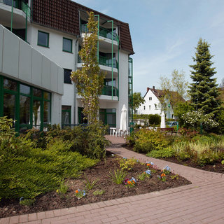 Weg durch einen Garten mit gepflegten Blumenbeeten, der zu einem modernen Wohngebäude führt. Mehrere Bäume und Sträucher umgeben den Weg, der von einer Terrasse mit Sonnenschirmen gesäumt ist. Im Hintergrund sind weitere Gebäude zu sehen.