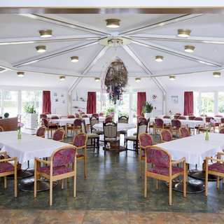 Ein hell erleuchteter Speisesaal mit runden Tischen, die mit weißen Tischdecken gedeckt sind. Stühle mit gepolsterten Rückenlehnen sind um die Tische gruppiert. An den Fenstern hängen rote Vorhänge, und es gibt einige Pflanzen im Raum, die eine einladende Atmosphäre schaffen.