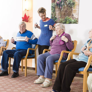Eine Gruppe älterer Menschen sitzt in einem Raum auf Stühlen. Eine Betreuerin leitet eine Bewegungsübung, während die Teilnehmer Hanteln halten und ihre Arme bewegen. Im Hintergrund ist ein Pflanzenmotiv zu sehen.