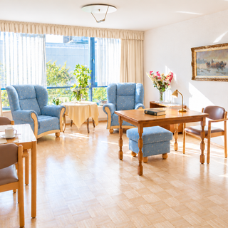 Ein helles und einladendes Wohnzimmer mit zwei blauen Sesseln und einem kleinen Tisch. Eine große Fensterfront lässt viel Tageslicht hinein, während ein Esstisch mit Obst und einer Flasche Wasser sowie eine Blumenvase auf dem Tisch stehen. Ein Schrank und ein Gemälde ergänzen den Raum.