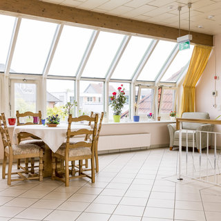 Der Raum ist hell und freundlich, mit großen Fenstern, die viel Tageslicht hereinlassen. In der Mitte steht ein Holztisch mit sechs Stühlen, umgeben von Pflanzen. Auf der rechten Seite befindet sich ein gemütlicher Sessel. Die Wände sind hell gestrichen und die Atmosphäre ist einladend.