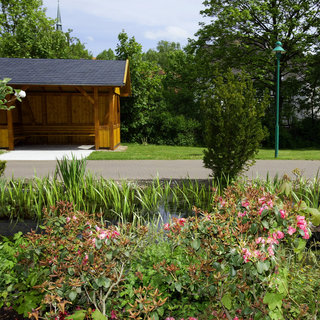 Ein Holzunterstand steht in einem grünen Park. Umgeben von üppigem Gras, Sträucher und Blumen mit rosa Blüten. Im Hintergrund sind Bäume und ein Weg zu sehen, der die Szenerie ergänzt.