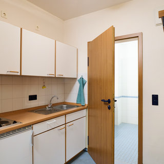 Eine kleine Küchenzeile mit weißen Schränken, einem Waschbecken und einer Kochplatte. Die Tür zur angrenzenden Nasszelle ist geöffnet. An der Wand hängt ein Handtuch, und neben der Tür befindet sich eine Kleiderablage.