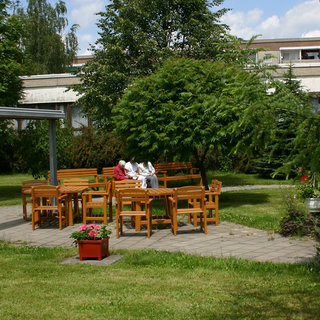 Eine ruhige Gartenszene mit mehreren Holzstühlen und -tischen unter einem großen Baum. Zwei Personen sitzen auf einer Bank, umgeben von üppigem Grün. Im Vordergrund steht ein blühender Pflanztopf. Der Himmel ist blau und voller Wolken.