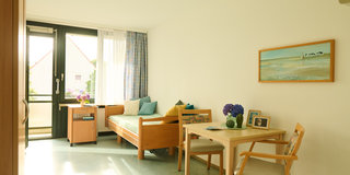 Ein gemütliches Zimmer mit einem Einzelbett, bunten Kissen und einem Tisch mit Stühlen. Am Fenster stehen blühende Pflanzen. An der Wand hängt ein Landschaftsbild. Der Raum ist hell und freundlich mit großen Fenstern, die viel Tageslicht hineinlassen.